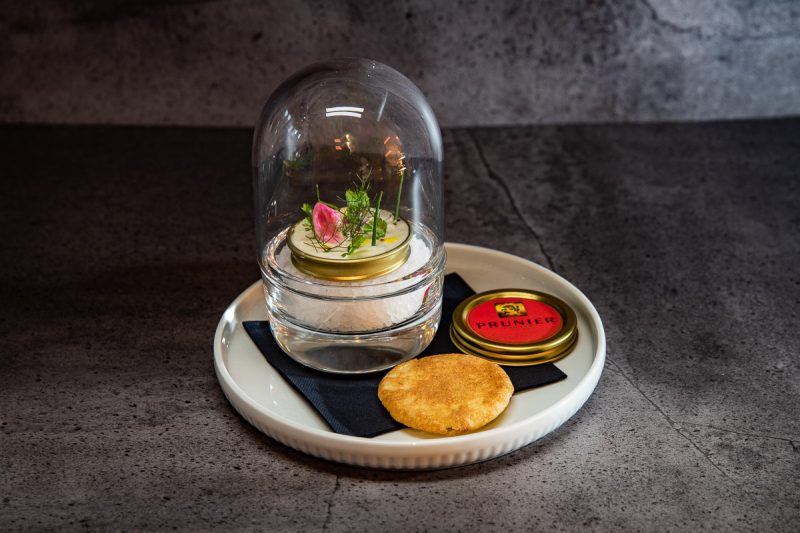 Caviarshot von Prunier mit Creme fraiche Haube und Kräutern unter einer Glasglocke mit Blini